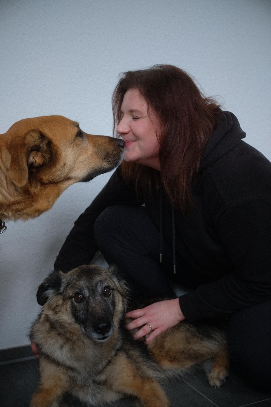 Kimberly Kralisch lächelt mit zwei Hunden — einer beschnuppert ihr Gesicht, der zweite liegt vor ihr.